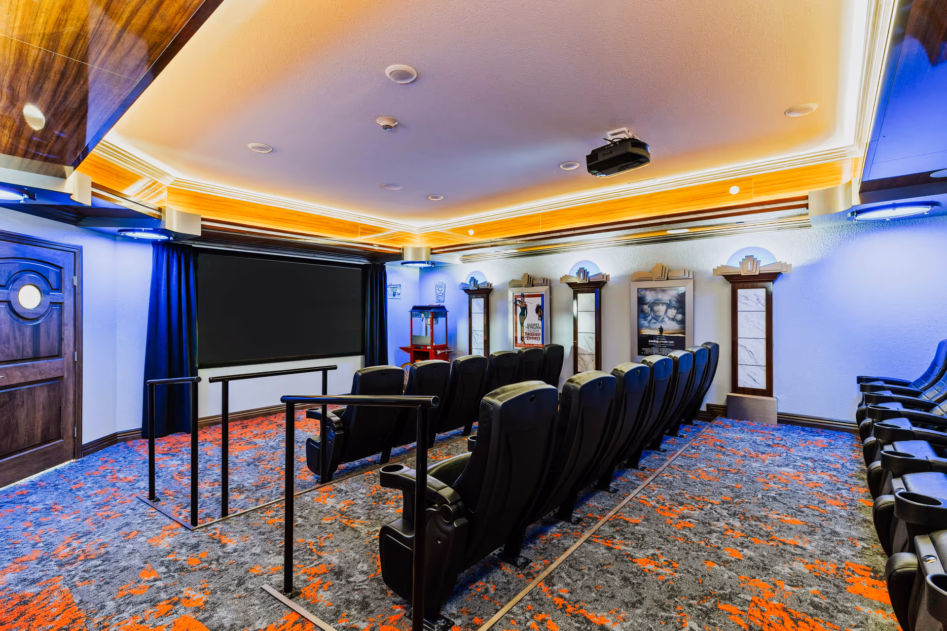 Interior view of a small movie theater room with rows of black reclining seats facing a large blank screen. The room features a patterned carpet with orange and gray colors, wood paneling on the ceiling with recessed lighting, and movie posters on the walls. There is a popcorn machine in the back corner and a wooden door on the left side.