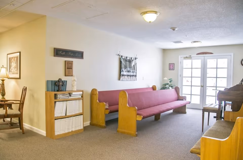 Interior room with wooden pew-style benches with red cushions, a piano with a bench, a small bookshelf with books and decorative items, a lamp on a side table, and double glass doors letting in natural light.