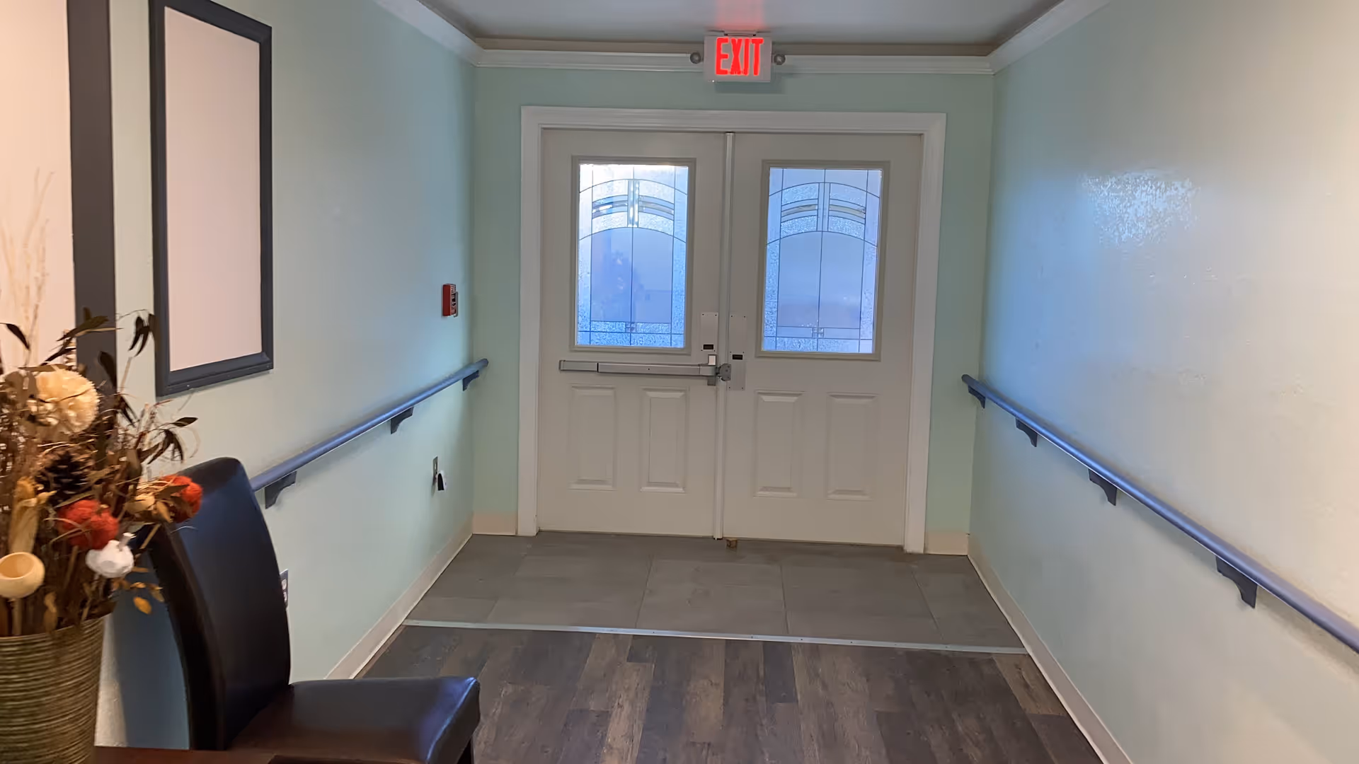 A hallway with light blue walls and wooden flooring leading to double doors with frosted glass panels. There are handrails on both sides of the hallway, a black chair and a vase with dried flowers on the left side, and an illuminated red exit sign above the doors.