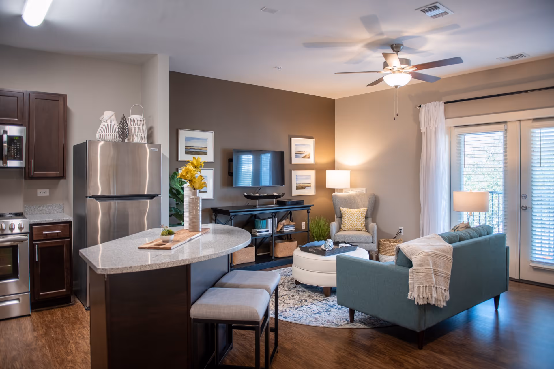 A modern open-concept living room and kitchen area featuring a stainless steel refrigerator, dark wood cabinets, a kitchen island with two stools, a blue sofa with a beige throw, a round ottoman, an armchair with a yellow patterned pillow, a TV mounted on a brown accent wall, framed pictures, and a ceiling fan with a light. French doors with white curtains lead outside.