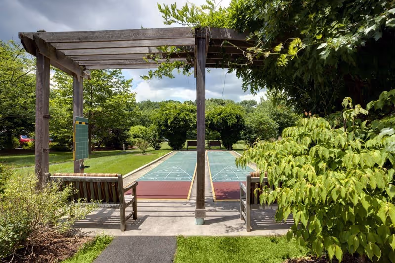 Outdoor shuffleboard court with two wooden benches under a wooden pergola surrounded by green trees and bushes on a sunny day with partly cloudy sky.