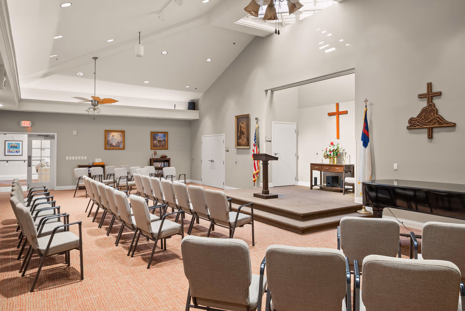 Interior view of a chapel or meeting room with rows of beige cushioned chairs facing a small stage with a wooden podium. The stage area has a wooden cross on the wall, a table with flowers, and flags. The room has a high ceiling with recessed lighting and ceiling fans, and religious artwork and symbols are displayed on the walls.