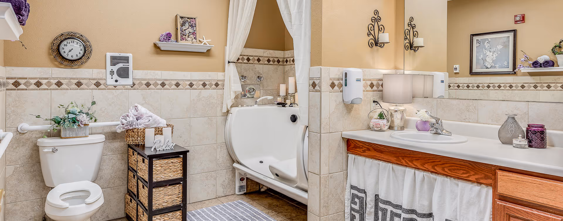 A well-decorated bathroom featuring a toilet with a flower arrangement on top, a wicker storage unit with towels and a 'Relax' sign, a walk-in bathtub with a curtain, and a sink with a large mirror above it. The walls are tiled halfway up with a decorative border, and the room has warm beige paint and wooden cabinetry.
