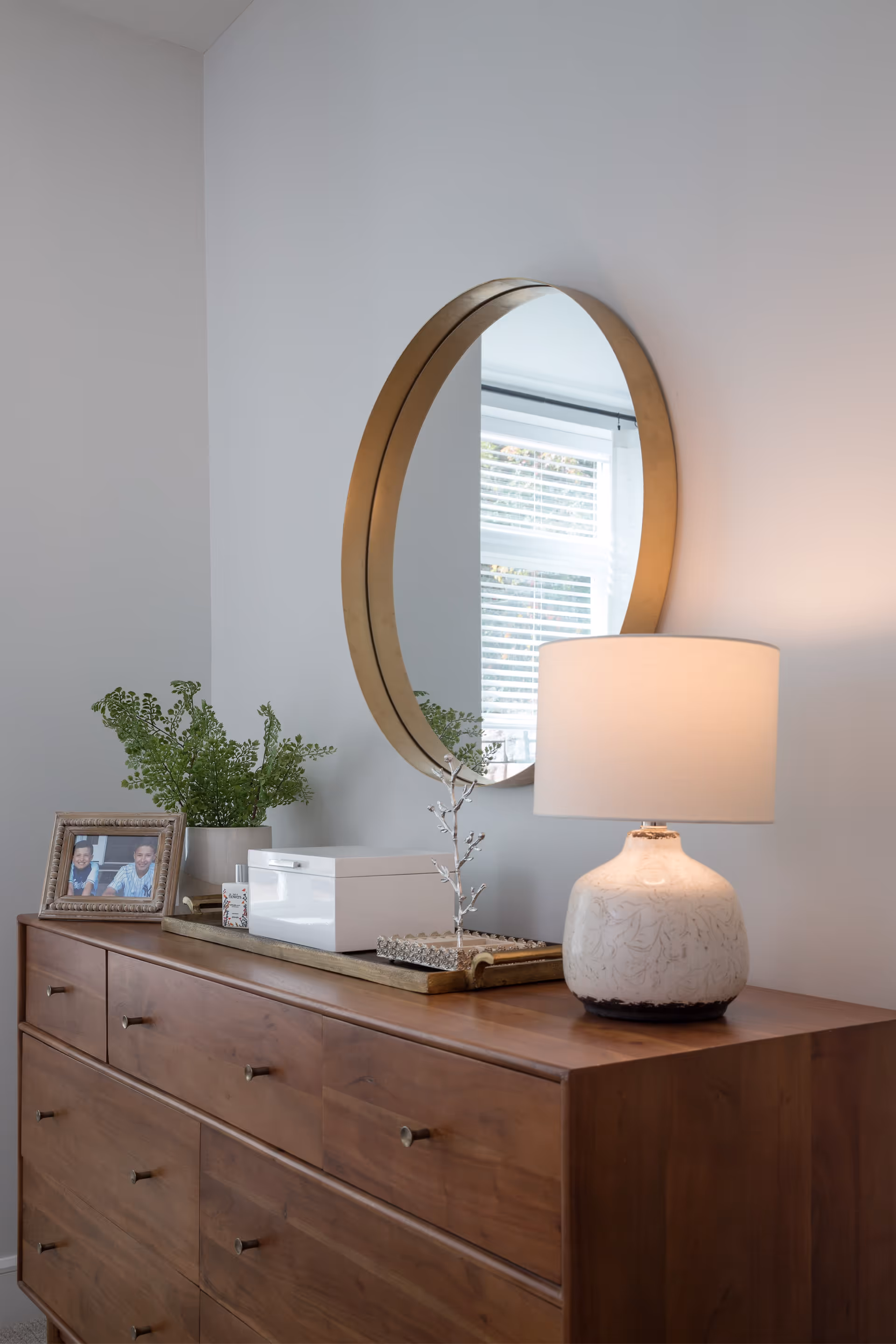 A wooden dresser with multiple drawers topped with a white decorative lamp, a round wall mirror with a gold frame, a small green plant in a pot, a framed photo, and a white box. The wall behind is light gray and a window with blinds is reflected in the mirror.