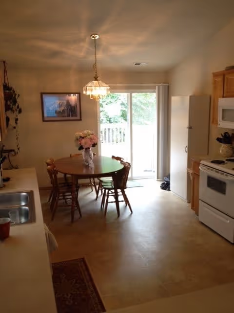 A kitchen and dining area with a round wooden table and four chairs in the center. A vase with pink flowers is on the table. To the right, there is a white stove, microwave, and wooden cabinets. On the left side, there is a countertop with a sink and a red cup. A sliding glass door at the back lets in natural light and shows greenery outside. A framed picture hangs on the wall above a small bench with plants nearby.