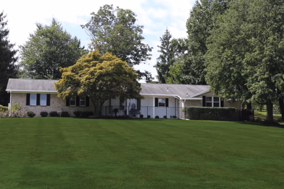 Single-story brick building with a gray roof surrounded by large green trees and a well-maintained grassy lawn in front under a partly cloudy sky.