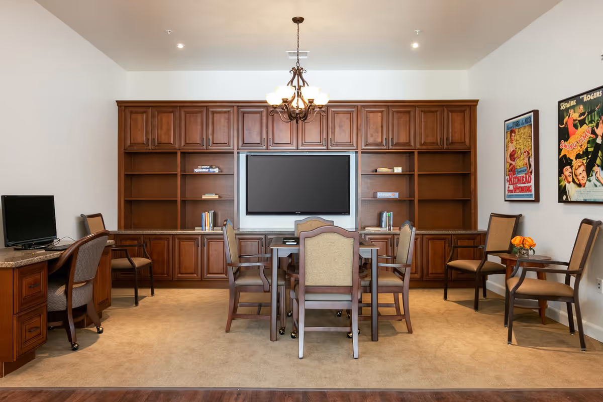 A well-lit common room with a large wooden built-in shelving unit and cabinets, a flat-screen TV mounted in the center, a table with six chairs in front of it, a desk with a computer on the left side, and two framed vintage posters on the right wall. The room has beige carpet and a chandelier hanging from the ceiling.