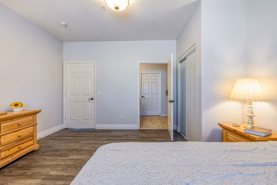 A bedroom with light-colored walls and wood flooring. There is a bed with a patterned bedspread in the foreground, a wooden dresser with a small sunflower decoration on the left, and a wooden nightstand with a lamp and a book on the right. Two white doors and a closet with sliding mirrored doors are visible on the far wall.