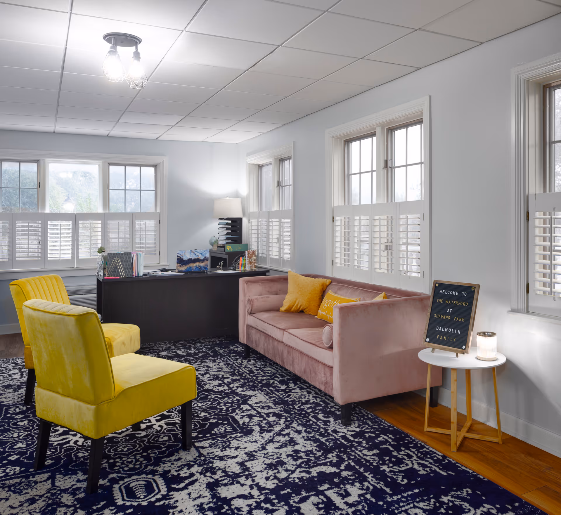 A bright and cozy office or reception area with a pink velvet sofa adorned with yellow pillows, two yellow upholstered chairs, a dark wooden desk with office supplies, a white side table with a candle and a welcome sign, and large windows with white shutters letting in natural light.