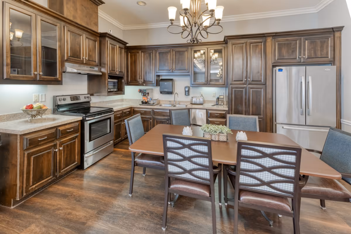 A spacious kitchen with dark wooden cabinets, stainless steel appliances including a stove and refrigerator, a wooden dining table with six chairs, and a chandelier overhead. The countertops are light-colored, and there is a small plant centerpiece on the table.