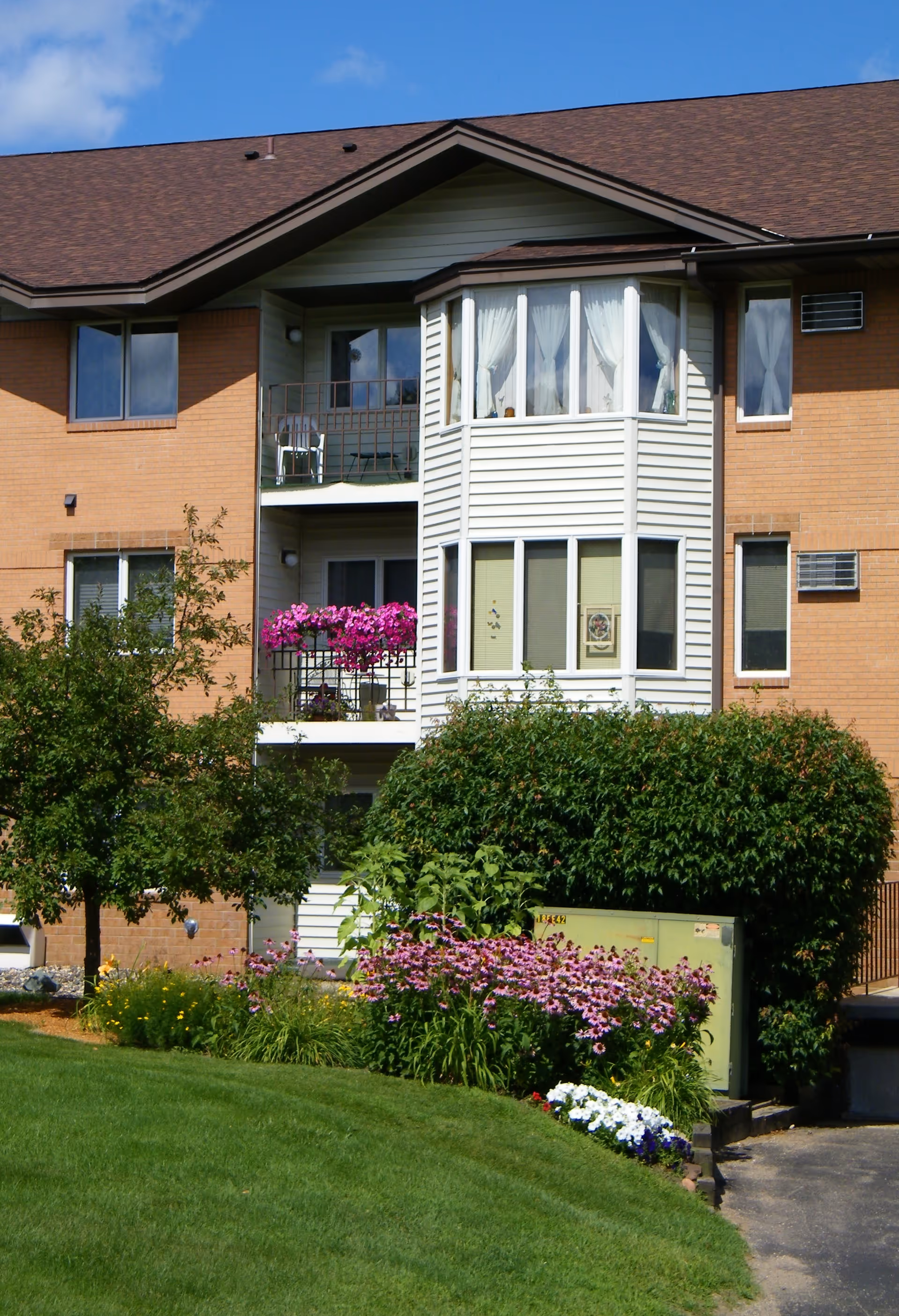 Exterior view of a senior living facility building with a brown roof and brick walls. The building features a balcony with pink flowers and a bay window with white curtains. There is a well-maintained green lawn and colorful flower beds in front of the building under a clear blue sky.