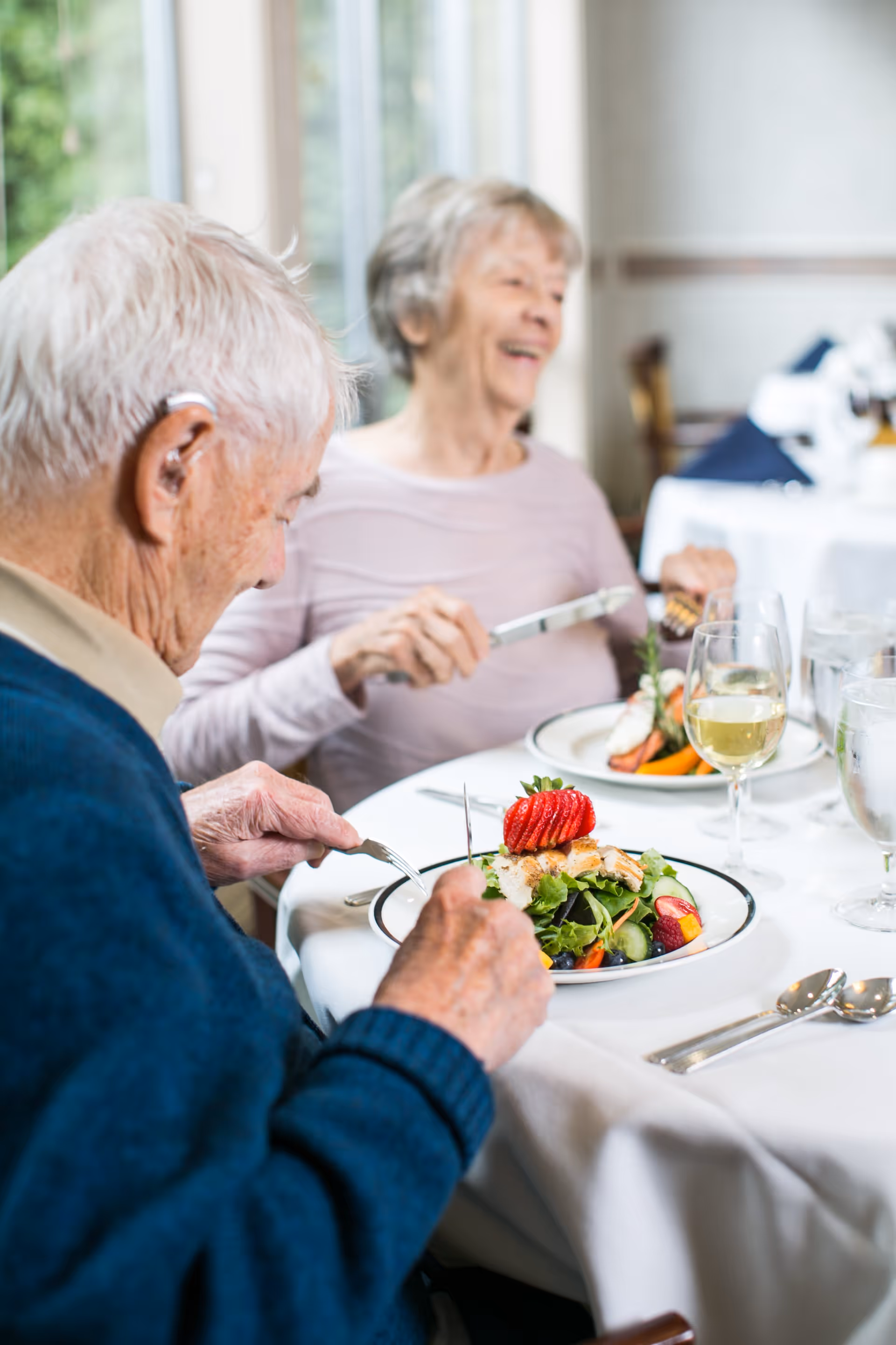 Two older adults seated at a white-clothed dining table enjoying plated salads and glasses of wine in a bright dining room.