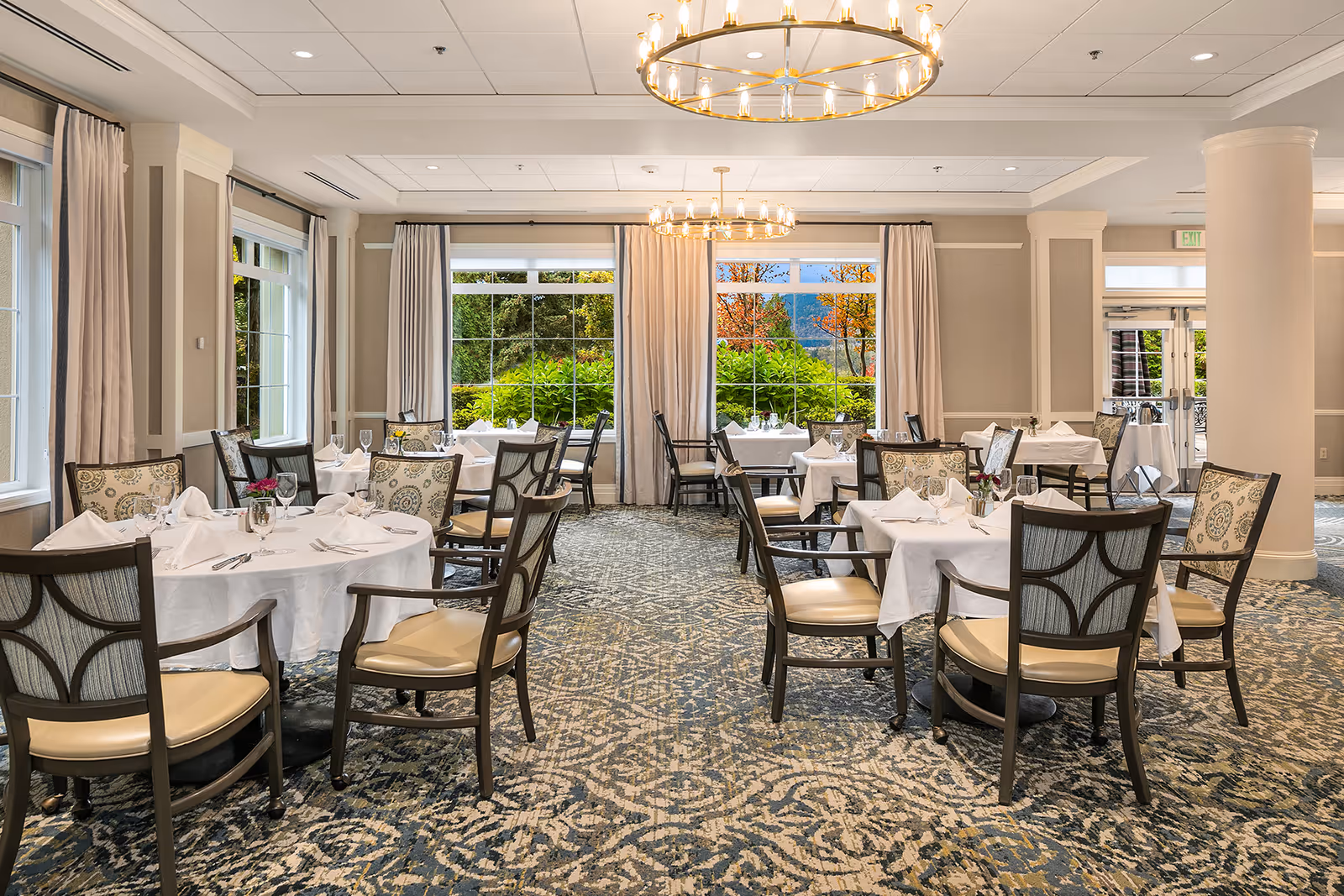 A bright and elegant dining room with round tables covered in white tablecloths, set with napkins, glasses, and silverware. The room features large windows with views of greenery and trees outside, beige walls, patterned carpet, and two circular chandeliers hanging from the ceiling.