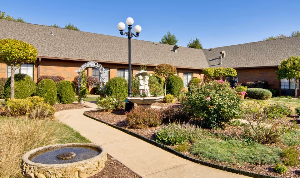 A landscaped garden courtyard with trimmed bushes, flowering plants, and a paved walkway. There is a decorative stone fountain with a statue of two children holding an umbrella, a black lamp post with four white globes, and a brick building with multiple windows in the background under a clear blue sky.