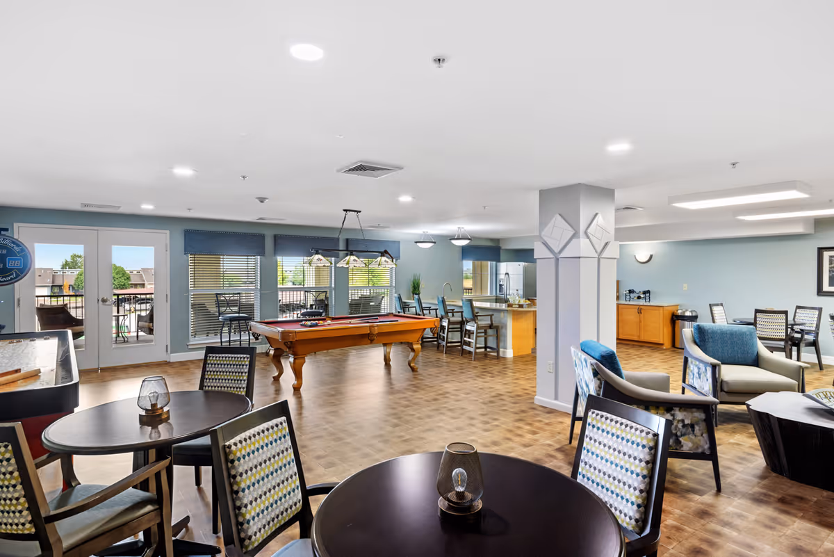A spacious senior living common area featuring a pool table in the center, several round tables with patterned chairs, a seating area with armchairs, and a kitchen area with bar stools. Large windows and glass doors provide natural light and access to an outdoor patio.