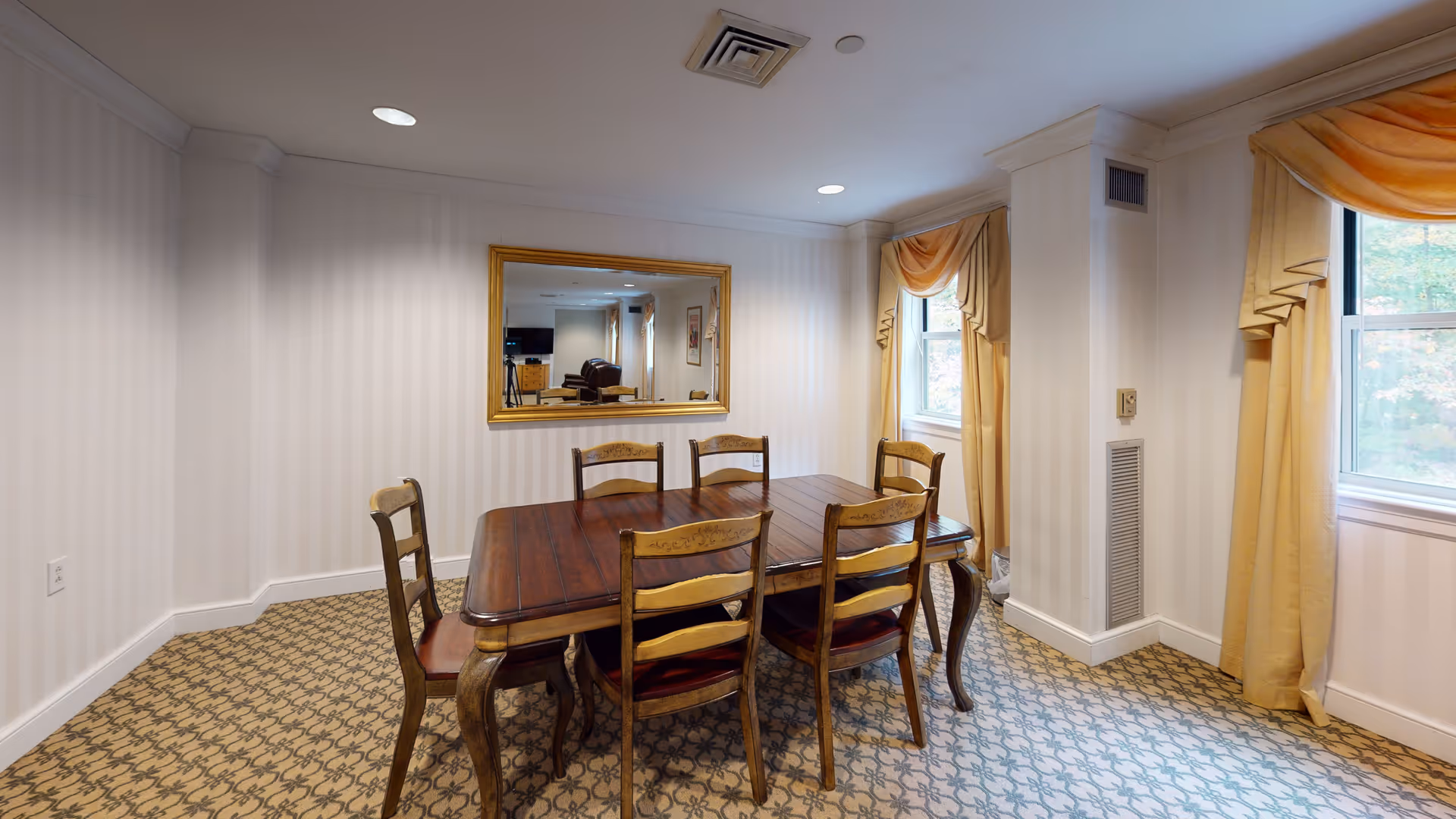 A dining room with a wooden table and six matching chairs. The room has patterned carpet, light-colored striped wallpaper, two windows with yellow curtains, and a large gold-framed mirror on the wall reflecting another room with a TV and furniture.