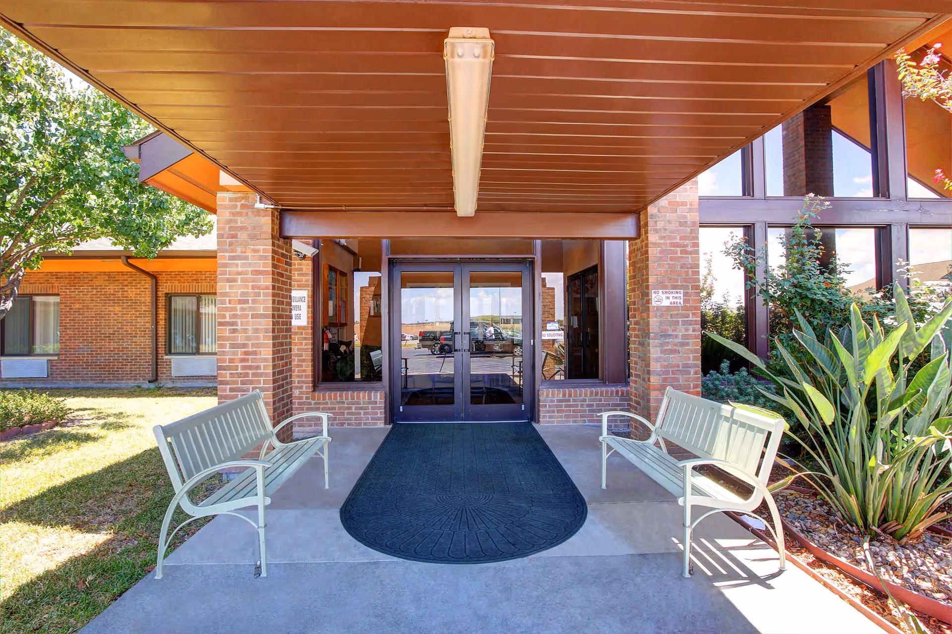 Entrance to Caraday of Corpus Christi Assisted Living facility with double glass doors, two white benches on either side of a black mat, brick walls, and plants on the right side under a brown overhang.
