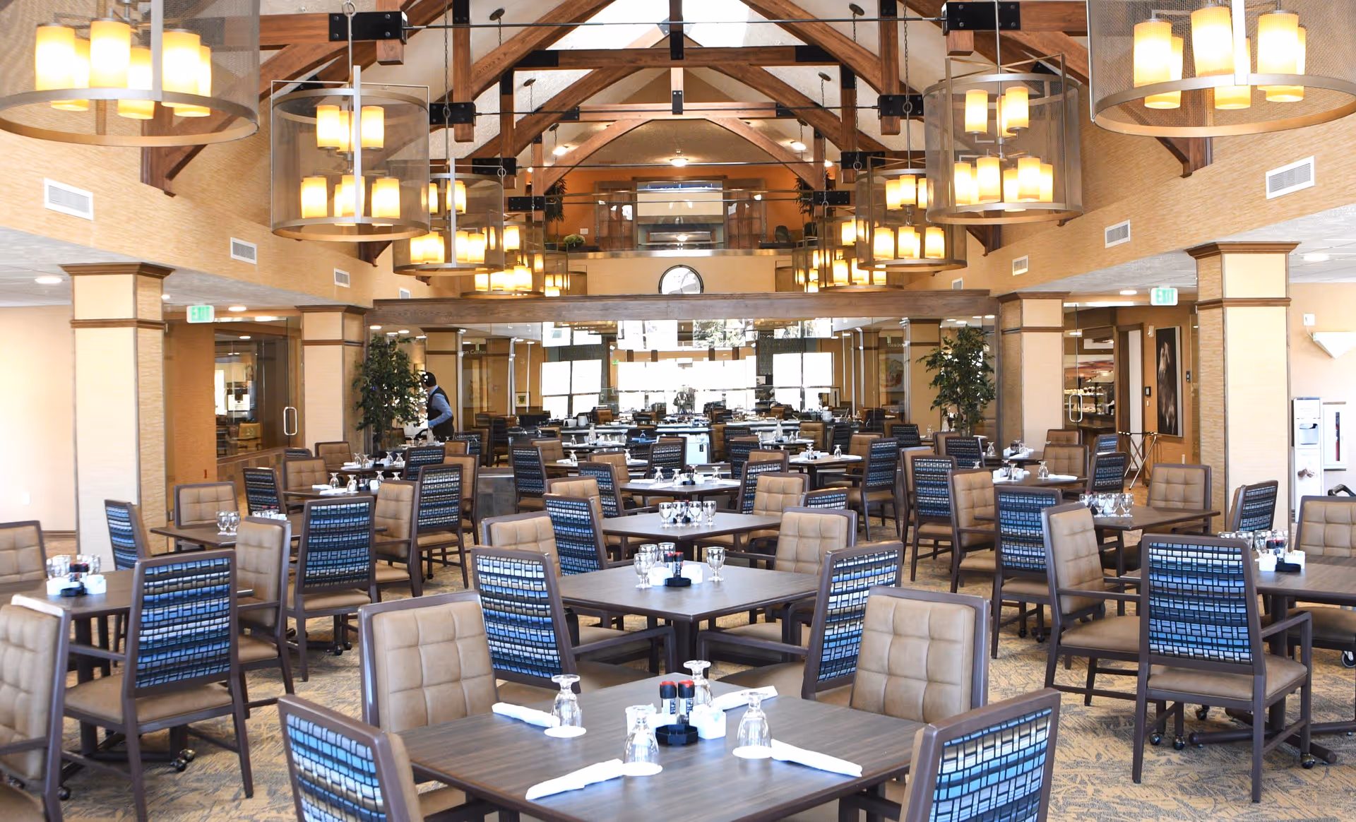 Spacious dining room with multiple tables and chairs arranged neatly. The room features high vaulted ceilings with wooden beams and several large hanging light fixtures. Tables are set with glasses, napkins, and condiments. There are plants and mirrors along the walls, creating a warm and inviting atmosphere.