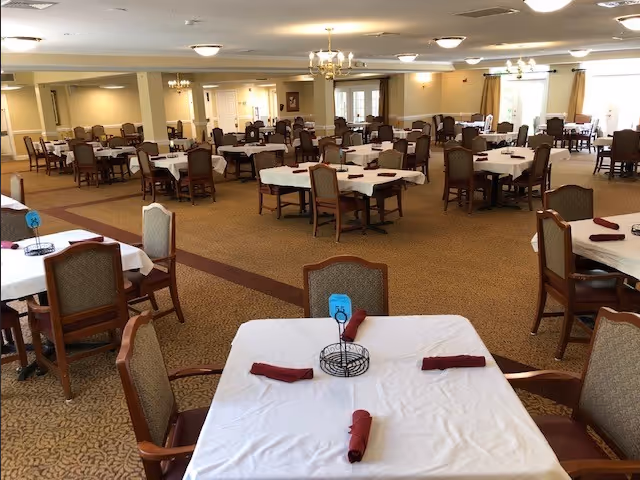 Large carpeted dining room with multiple tables covered in white tablecloths, chairs, chandeliers, and rolled napkins.