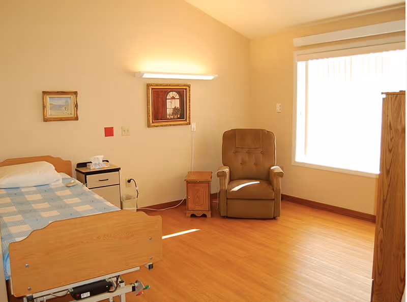 A simple, well-lit bedroom in a senior living facility with a single bed covered by a blue and white checkered blanket, a small bedside table with tissues, a brown cushioned armchair, a wooden side table, two framed pictures on the wall, and a large window with vertical blinds letting in natural light.