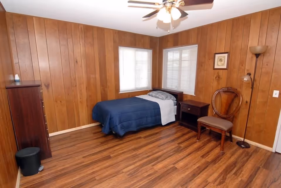 A simple bedroom with wood-paneled walls and wood flooring. The room contains a single bed with a blue comforter and white sheet, a wooden nightstand, a wooden dresser, a wooden chair with a cushioned seat, a floor lamp, and two windows with blinds.