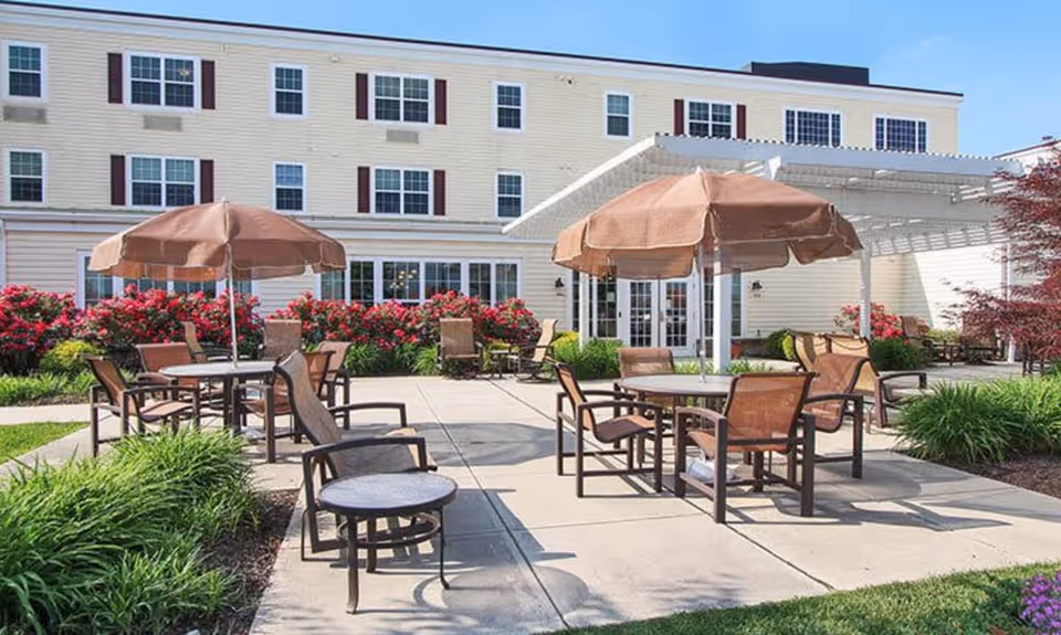 Outdoor patio area at Keystone Villa at Fleetwood featuring several round tables with brown umbrellas and matching chairs. The patio is surrounded by well-maintained landscaping including green bushes and red flowering plants. The building's light yellow exterior with multiple windows is visible in the background under a clear blue sky.