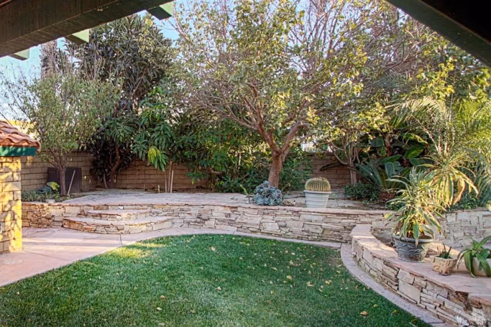 A landscaped outdoor garden area with a curved stone retaining wall, green grass, various potted plants, and trees. The area is partially shaded by a roof structure.