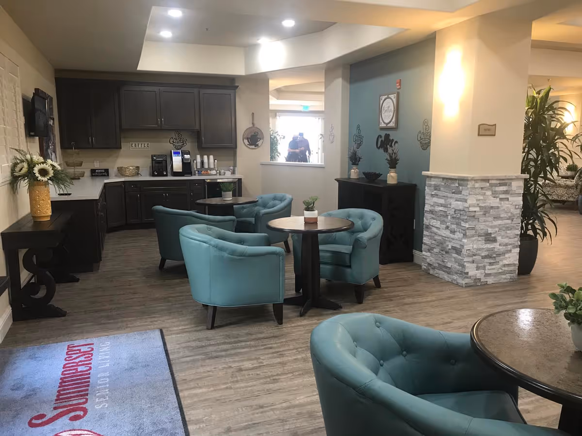 Bright senior living lounge with teal armchairs clustered around small round tables and a coffee station in the background.