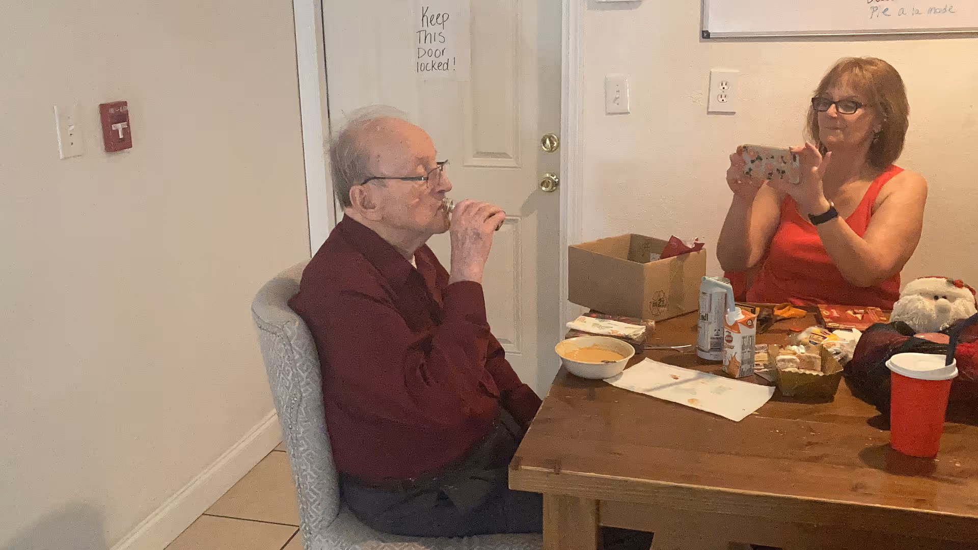 An elderly man wearing glasses and a maroon shirt is sitting at a wooden table drinking from a small bottle. Across from him, a woman in a red sleeveless top is holding a smartphone, seemingly taking a photo or video. The table has various items on it including a bowl, cans, snacks, and a red cup. The setting appears to be a casual indoor room with a white door and light-colored walls.
