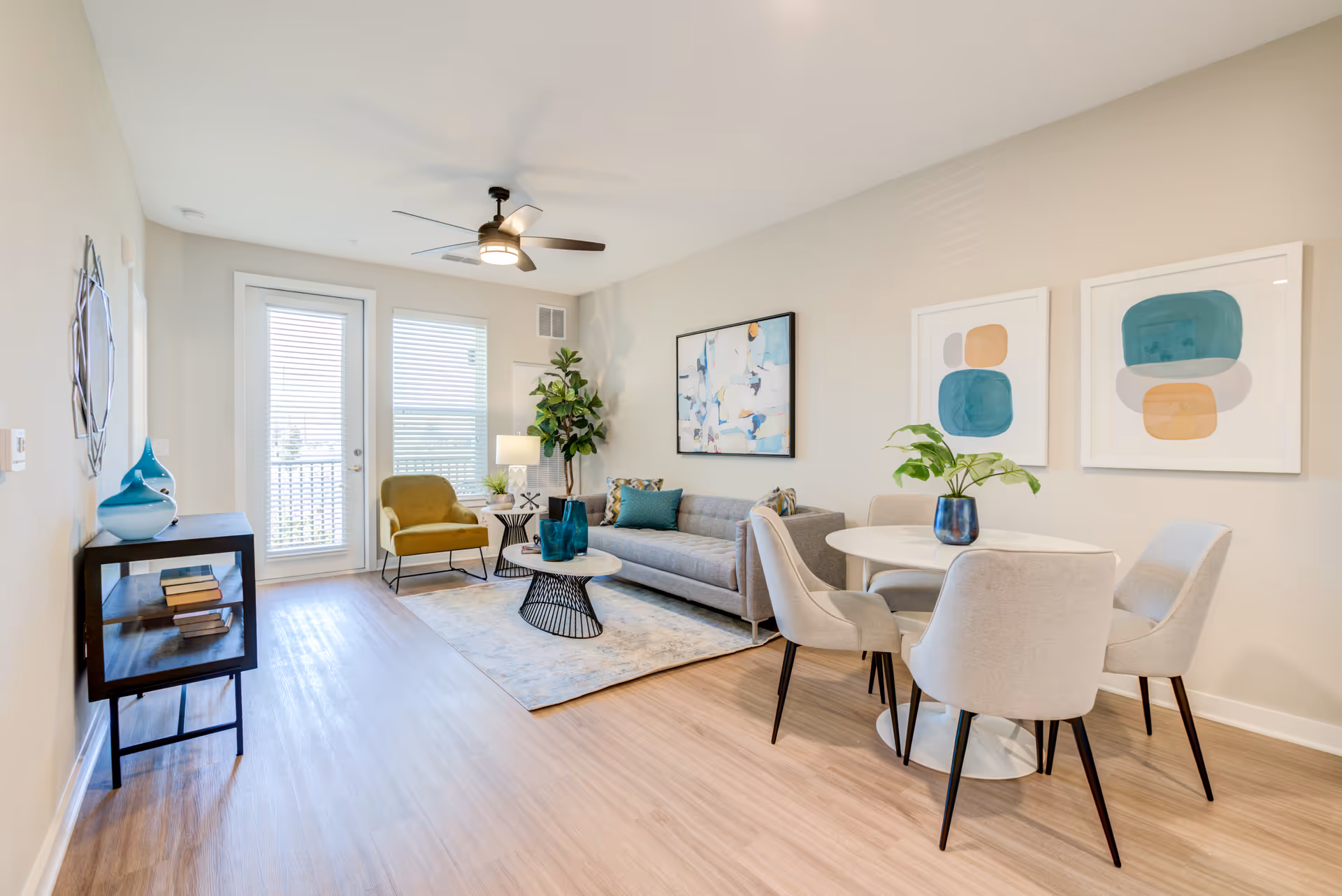 A bright and modern living room and dining area with light wood flooring. The living room features a gray sofa with blue and patterned cushions, a round coffee table with blue decorative vases, a mustard yellow armchair, a side table with a lamp, and a tall green plant. The dining area has a white round table with four light gray upholstered chairs and a blue vase with green leaves. The walls are decorated with abstract artwork in blue, beige, and gray tones. A ceiling fan with a light is mounted on the white ceiling, and there are large windows with blinds and a glass door letting in natural light.