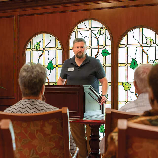 A man stands behind a wooden podium speaking to an audience seated in chairs. Behind him are three large stained glass windows with a leafy design. The room has wooden paneling and the audience members are partially visible from behind.