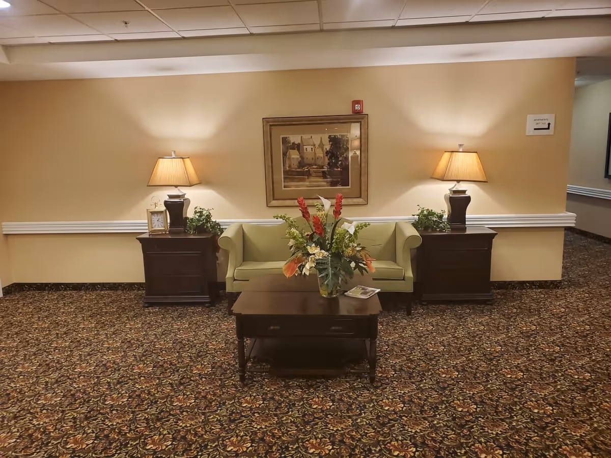 A cozy seating area in a senior living facility hallway featuring a light green loveseat flanked by two dark wooden side tables with lamps and plants. A dark wooden coffee table with a floral arrangement and a magazine is placed in front of the loveseat. A framed painting hangs on the beige wall behind the loveseat.