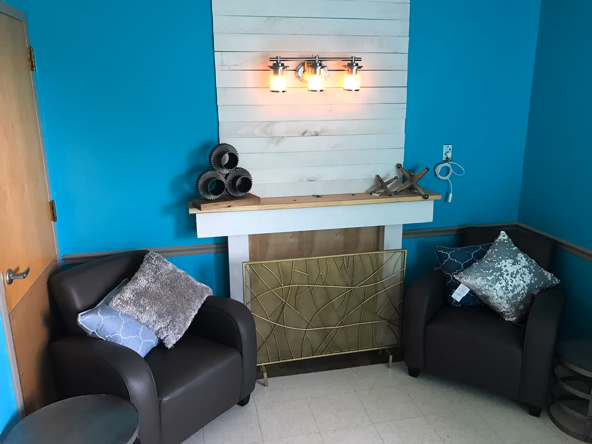 A cozy corner of a room with two dark brown armchairs, each adorned with decorative pillows. Between the chairs is a white fireplace with a decorative metal screen in front. The wall behind the fireplace is painted turquoise, and there is a three-light wall sconce above the fireplace. Decorative items are placed on the mantel, and a wooden door is visible to the left.