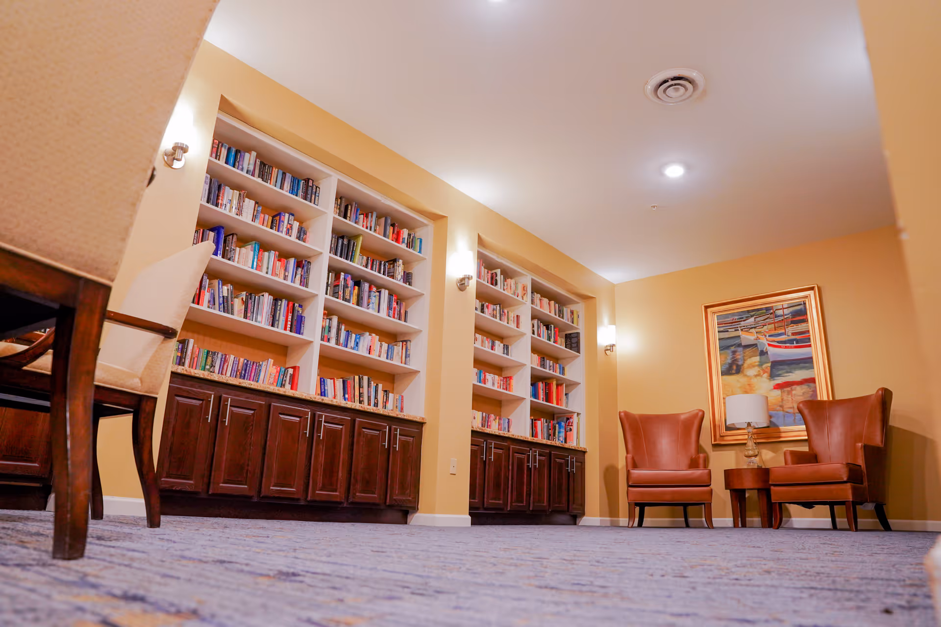 A cozy reading area in a senior living facility featuring built-in bookshelves filled with books, two brown leather armchairs, a small wooden side table with a lamp, and a framed painting of boats on the wall. The room has warm yellow walls and carpeted flooring.