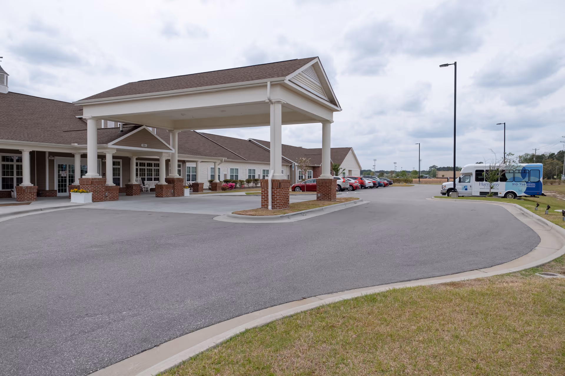 Exterior view of Harmony at Hope Mills facility showing the main entrance with a covered drop-off area supported by white columns with brick bases. Several cars are parked in the parking lot to the right, and a shuttle bus with the Harmony logo is visible. The sky is cloudy.
