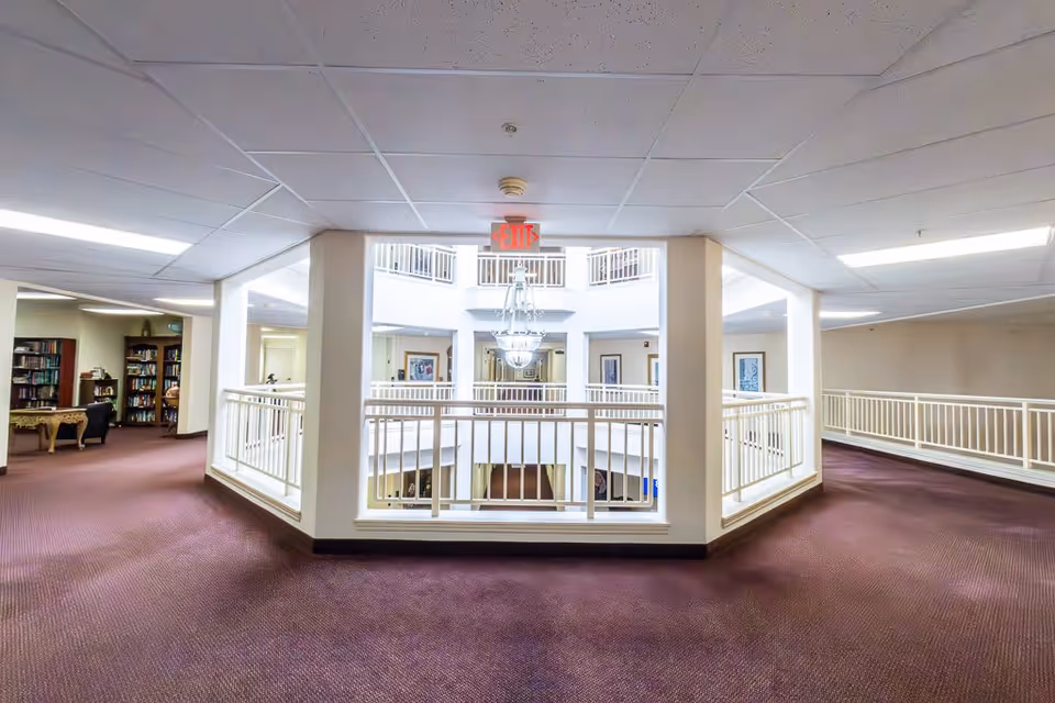 Interior view of a senior living facility hallway with maroon carpet, white railings overlooking a central atrium, and a chandelier hanging in the middle. There are bookshelves and seating areas visible on the left side.