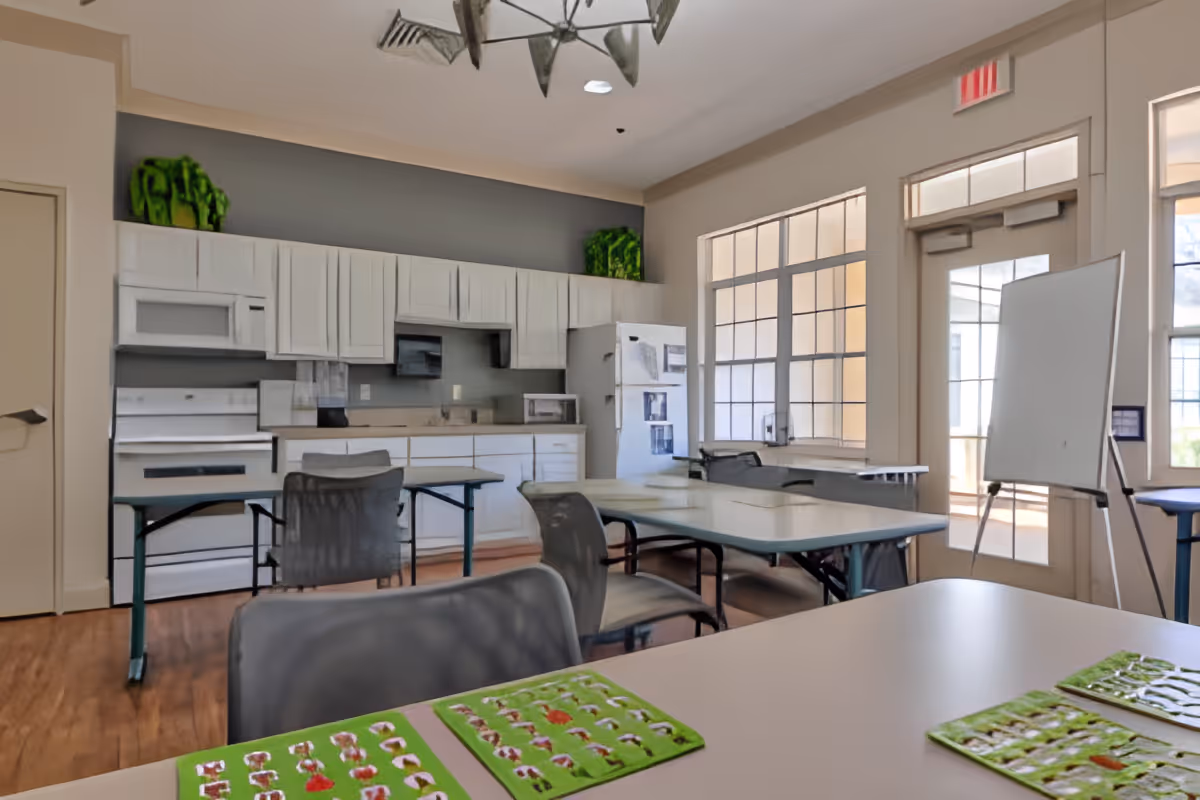 A bright kitchen and dining area with white cabinets, a white stove, microwave, and refrigerator. Several tables and chairs are arranged in the room, with a whiteboard on an easel near large windows and a glass door letting in natural light. Green decorative plants are placed on top of the cabinets.