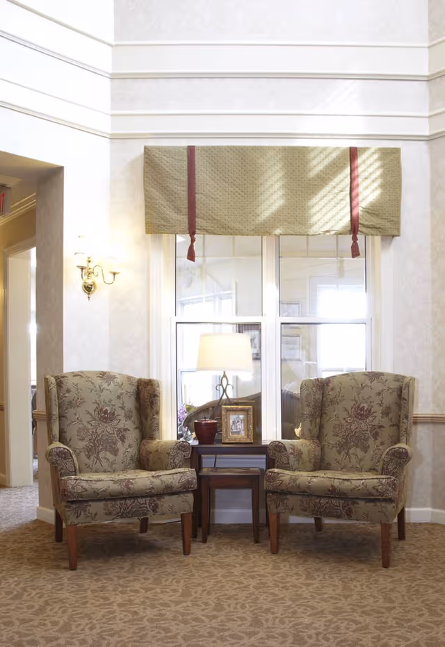 Two patterned armchairs with floral designs are positioned on either side of a small wooden table with a lamp, a framed photo, and a decorative item. Behind the chairs is a window with a green valance with red tassels. The room has beige patterned carpet and wallpaper, with a wall sconce light on the left side.