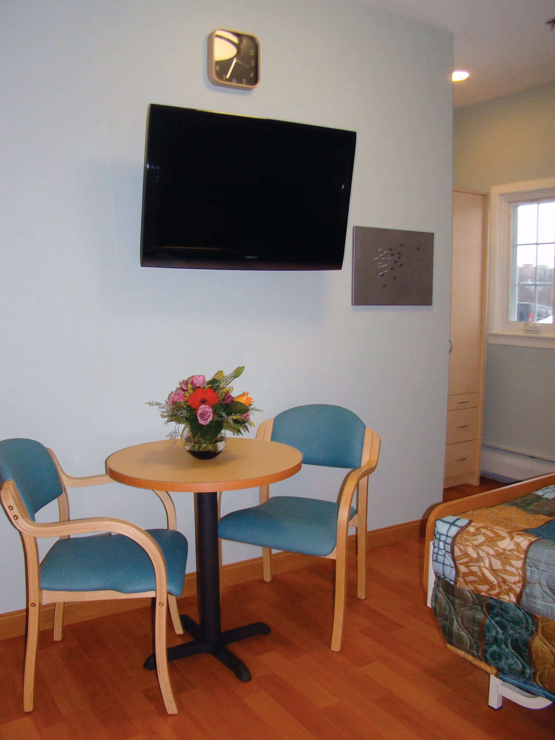 Small room with two teal chairs and a round table holding a flower arrangement, a wall-mounted TV and clock, and part of a bed visible.
