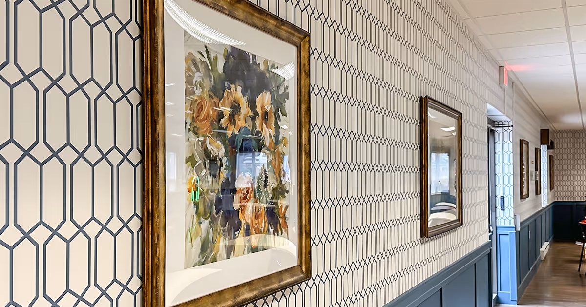 A hallway with patterned wallpaper and framed floral artwork on the walls. The lower half of the walls is painted blue with wainscoting. The hallway has a wooden floor and a ceiling with recessed lighting and an exit sign visible in the distance.