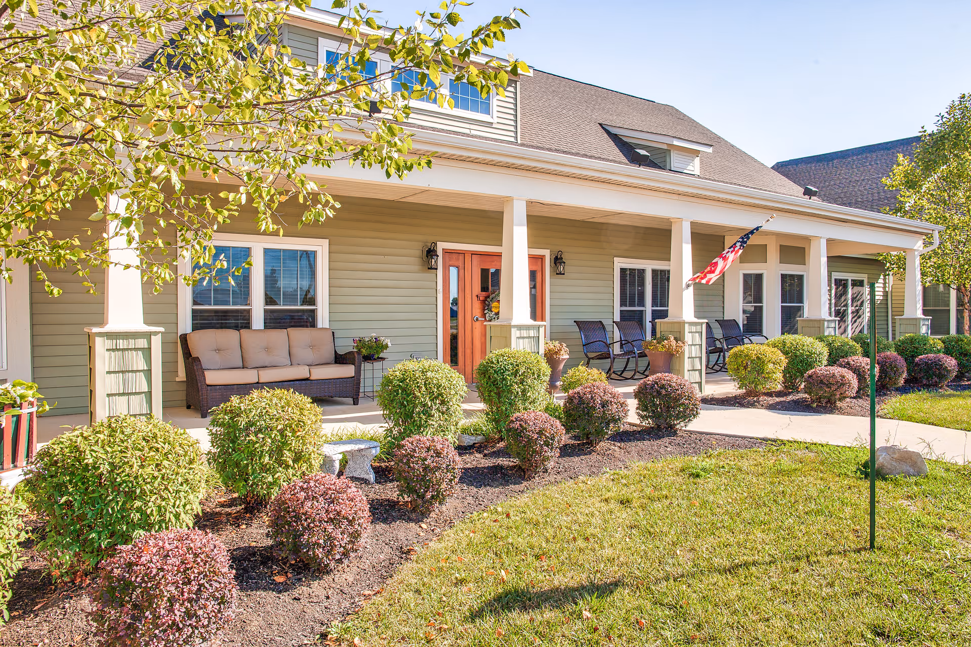 Front porch of a senior living facility with a beige cushioned sofa, several rocking chairs, green and purple shrubs, an American flag, and a well-maintained lawn under a clear blue sky.