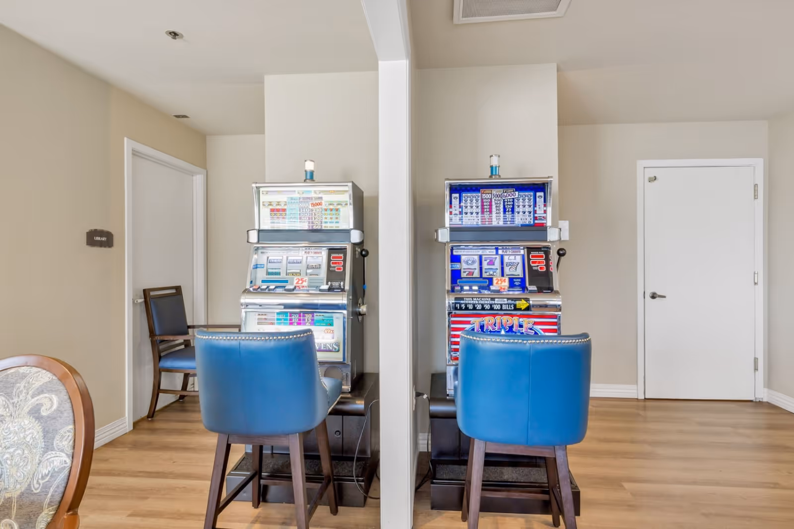 Two vintage slot machines placed side by side against a wall with a white partition between them. Each machine has a blue chair in front of it. The room has light-colored walls, wooden flooring, and two closed white doors, one with a sign labeled 'Library'.