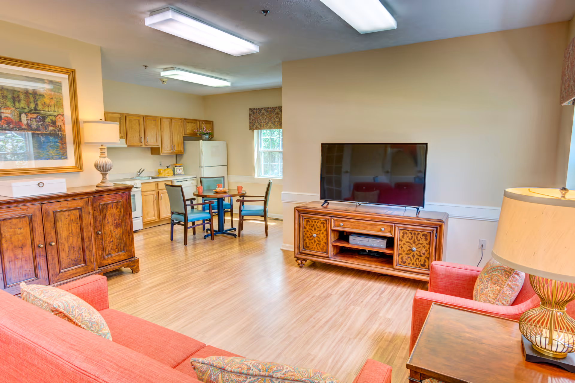 A bright and cozy living area with a red sofa and armchair, a wooden TV stand with a flat-screen television, a small dining table with four chairs, and a kitchen area with wooden cabinets, a refrigerator, and a stove. The room has wooden flooring, beige walls, and is well-lit with ceiling lights and table lamps.