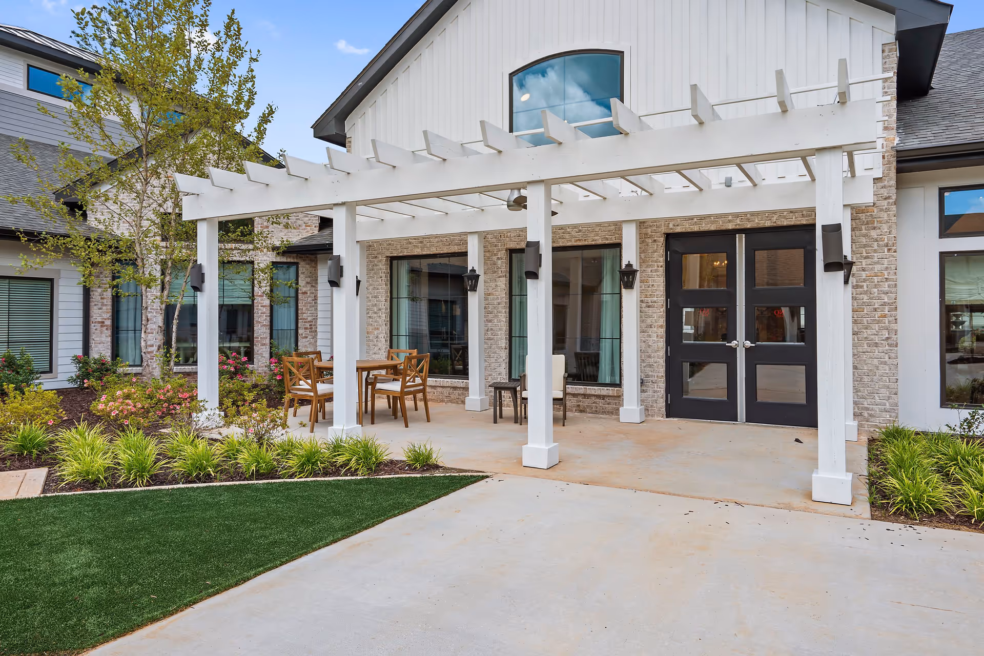 Outdoor patio area at Sage Lake facility featuring a white pergola with wooden chairs and tables underneath, surrounded by landscaped greenery and flowers, with large windows and double black doors on the building facade.
