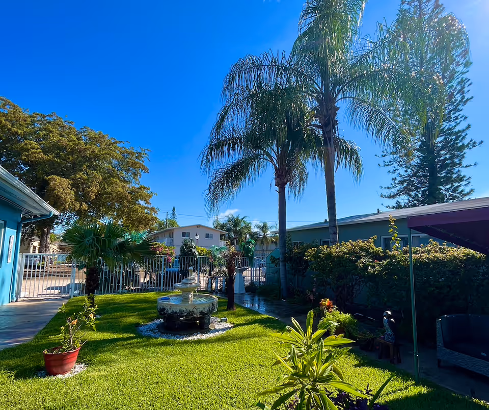 Sunny outdoor courtyard with palm trees, a fountain, blooming plants, and surrounding buildings.