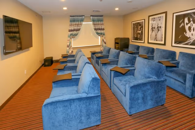 A small theater room with two rows of blue upholstered reclining chairs, each equipped with a small black tray table. The room has a striped carpet, beige walls, a flat-screen TV mounted on the wall, and three black and white framed pictures on the right wall. A window with patterned curtains is at the back of the room.