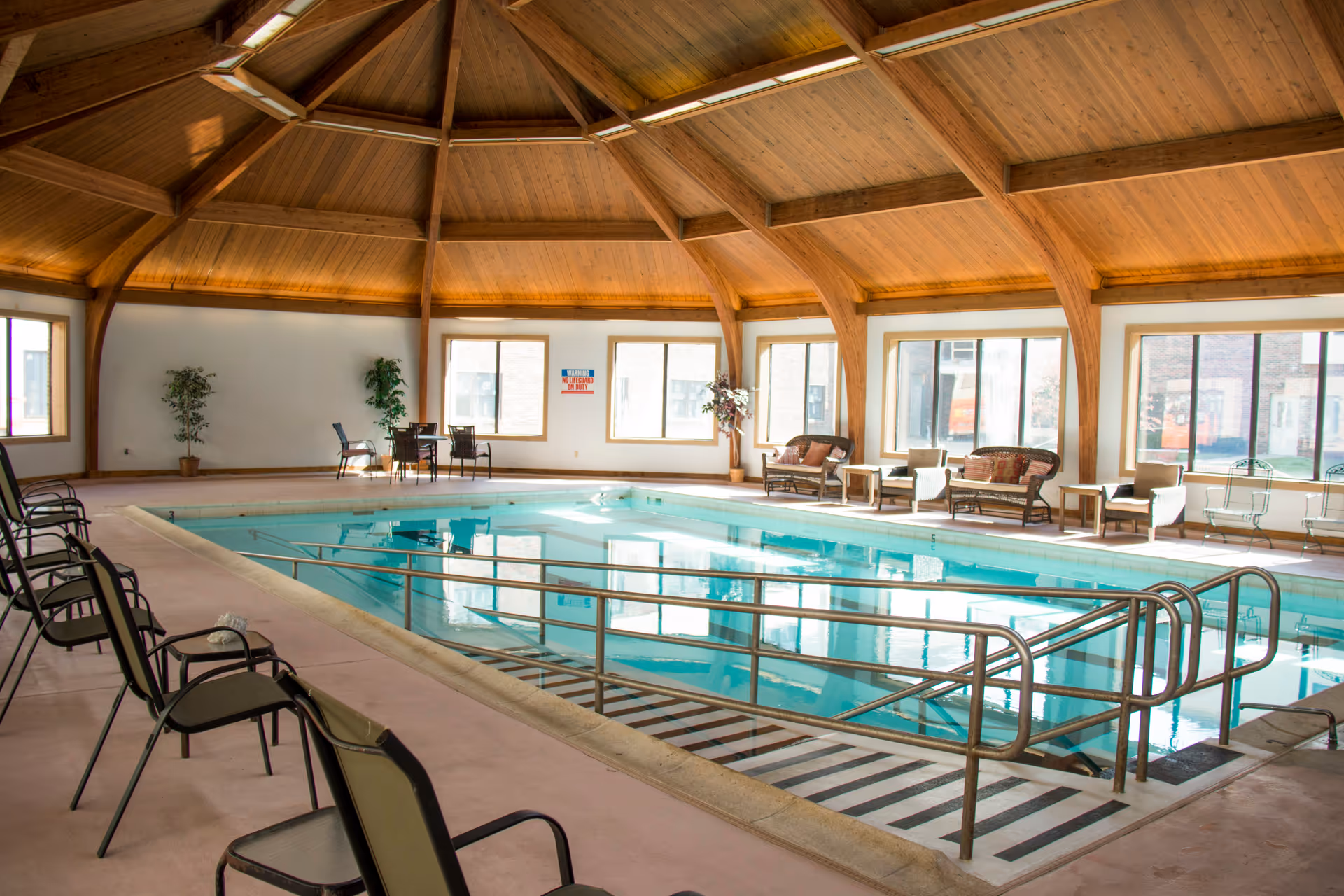 Indoor swimming pool with a wooden vaulted ceiling and large windows letting in natural light. The pool has metal handrails and a ramp for accessibility. Around the pool are several chairs and small tables, along with some potted plants.