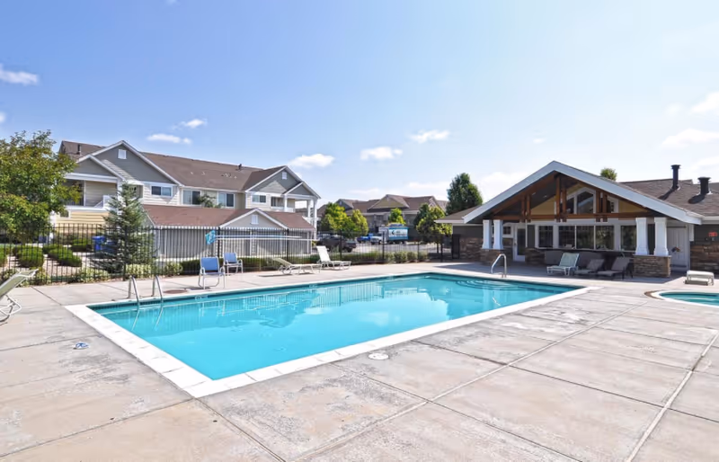Outdoor swimming pool and patio with lounge chairs in front of a clubhouse and nearby residential buildings under a clear sky.