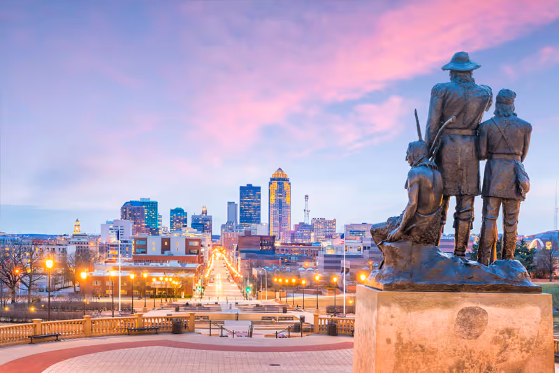 View of a city skyline at dusk with a pink and blue sky, seen from a raised platform with a bronze statue of three historical figures in the foreground.