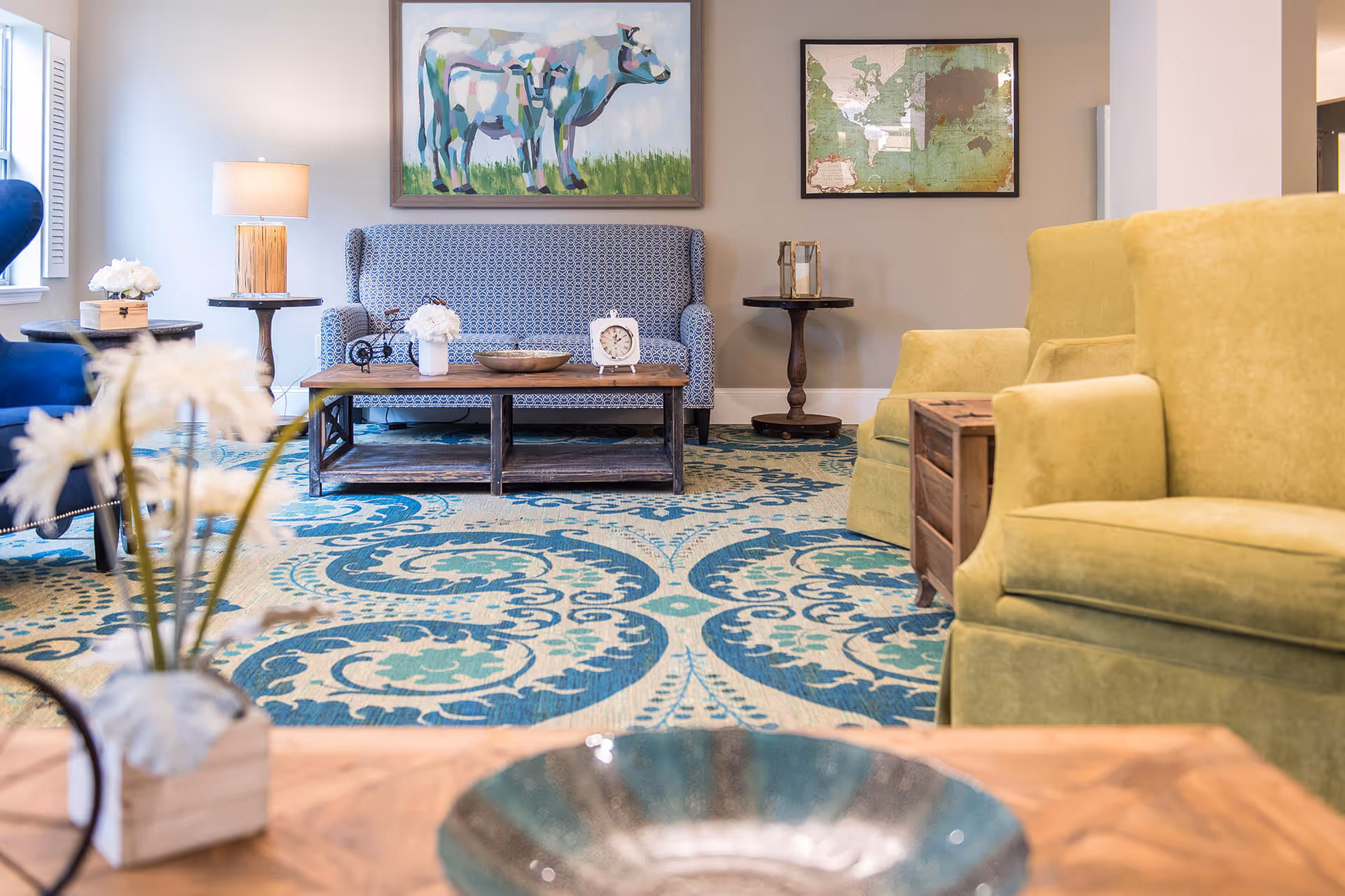 A cozy living room area with a patterned blue loveseat, two green armchairs, a wooden coffee table with decorative items including a clock and flowers, a blue patterned rug, and two framed artworks on the wall, one depicting a cow and the other a world map.